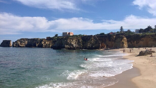 Une plage côtière aux eaux cristallines, bordée de falaises rocheuses et sur laquelle se trouve un bâtiment perché au sommet de la falaise.