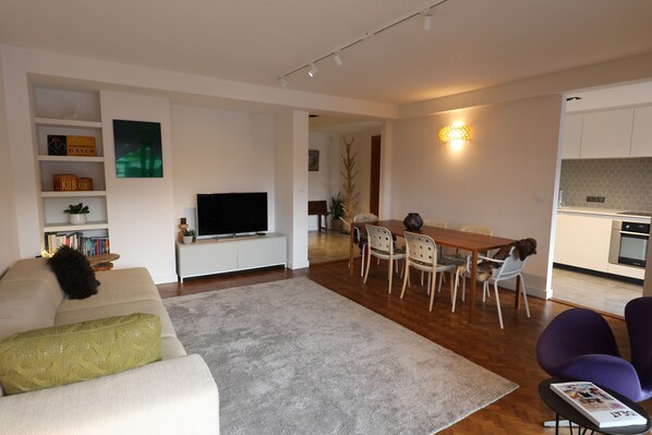A modern living room with a flat-screen TV, a dining area with a wooden table and chairs, and a kitchen in the background.