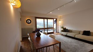 A modern living room with a wooden dining table, a sofa, and a view of a mountain landscape through large windows.
