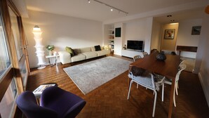 A modern living room with a wooden floor, a sofa, a dining table with chairs, a TV, and a bookshelf.
