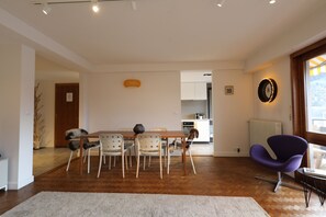Een eetgedeelte met een houten tafel en stoelen, een keuken op de achtergrond en een paarse fauteuil.