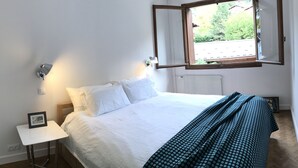 A neatly made bed with white bedding and a green patterned throw. There is a small bedside table with a framed picture. A window with a wooden frame is visible, showing greenery outside.
