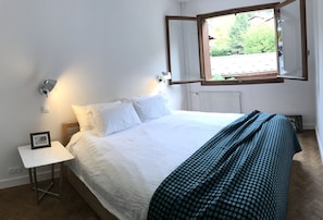 Una cama bien tendida con ropa blanca y un plaid verde estampado. Hay una mesita de noche pequeña con un cuadro enmarcado. Se ve una ventana con marco de madera, mostrando follaje en el exterior.