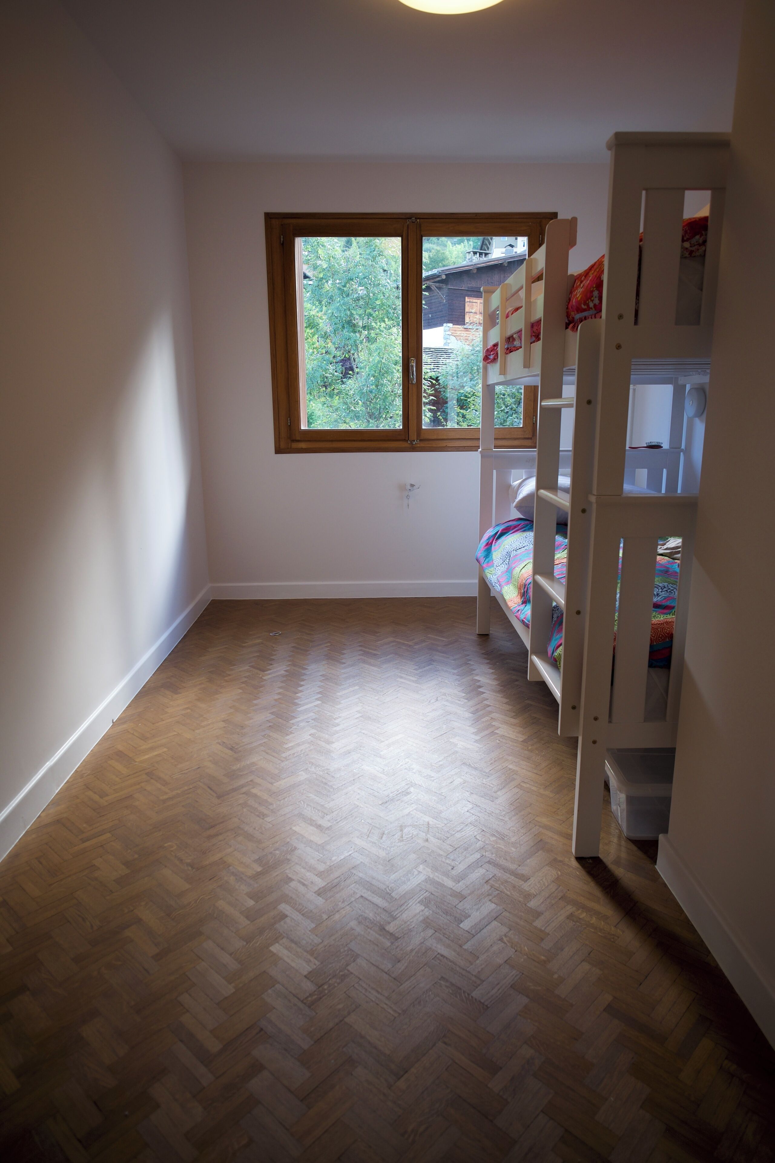 A room with a bunk bed, a window with a view of greenery, and a herringbone wooden floor.