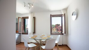 A dining area with a glass table, white chairs, and a view of buildings through a window. - Venice Appartament Salini II (Venezia)