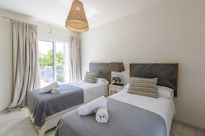 A hotel room with two beds, a large window with curtains, a hanging light fixture, and a nightstand with a lamp and a decorative elephant.