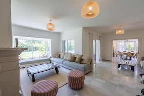 A spacious living room with a sofa, ottomans, and a coffee table. There are large windows and a dining area visible in the background.