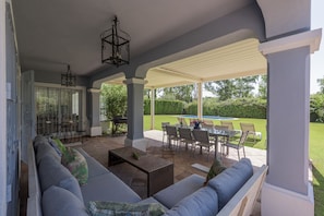 A covered patio area with a seating arrangement, a table, and a view of a swimming pool and garden.