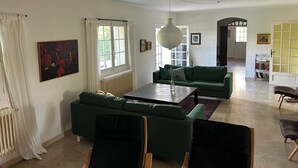A spacious living room with a green sofa set, a coffee table, and a hanging light fixture. There is a radiator on the wall and a door leading to another room.