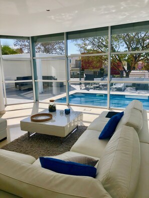 A modern living room with a large glass wall overlooking a swimming pool, a white sofa with blue cushions, and a coffee table with decorative items.