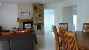 Un salon avec une cheminée en pierre, un canapé gris et un coin repas avec des chaises et une table en bois.