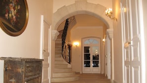 Um amplo corredor com piso de pedra, um banco de madeira, escada com corrimão de ferro forjado e uma porta com painéis de vidro.