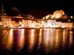 A nighttime coastal town with a prominent castle on a hill, boats docked along the waterfront, and illuminated buildings.