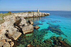 A rocky coastline with clear blue waters, a lighthouse, and a distant view of the sea.