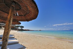 Een strand met rieten parasols en witte ligstoelen.