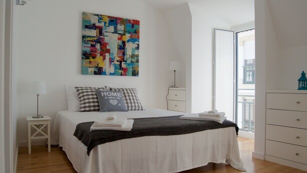 A bedroom with a large bed, a colorful abstract painting, a nightstand with a lamp, and a view of the outside through an open door. - House in the Historic Center of Cascais (Cascais)