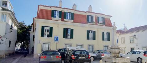 Una calle empedrada con coches aparcados y un edificio con tejado rojo y contraventanas verdes.