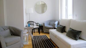 A modern living room with a white sofa, a grey armchair, a round mirror, a small table with a chair, and a window providing natural light. - House in the Historic Center of Cascais (Cascais)