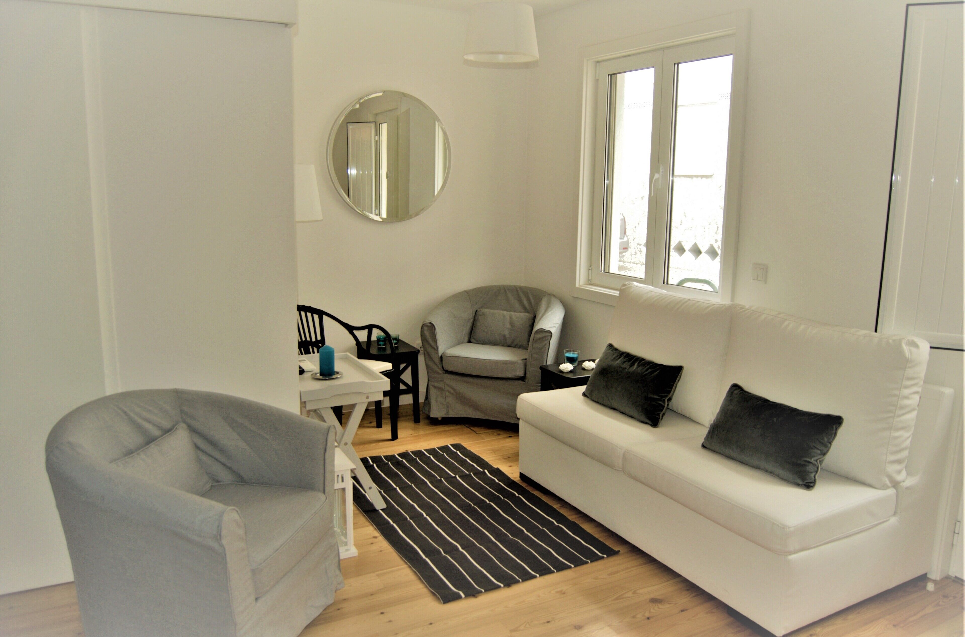 A modern living room with a grey armchair, a white sofa with black cushions, a round mirror, a small table with a lamp, and a window with a view.