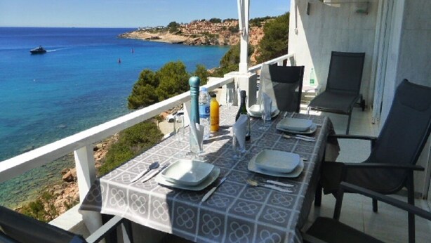 Un balcon avec une table dressée pour le dîner, offrant une vue sur une zone côtière aux eaux d’un bleu limpide et sur une côte lointaine.