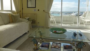 Un salon avec un canapé, une table basse en verre sur laquelle sont posés des magazines, un tableau encadré au mur et une vue sur le port de plaisance, visible à travers de grandes fenêtres.