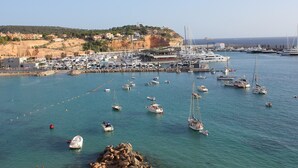 Un port de plaisance où sont amarrés de nombreux bateaux, une côte rocheuse et une colline avec des bâtiments en arrière-plan.