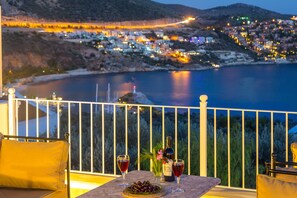 A balcony with a table set for two, overlooking a coastal town at dusk.