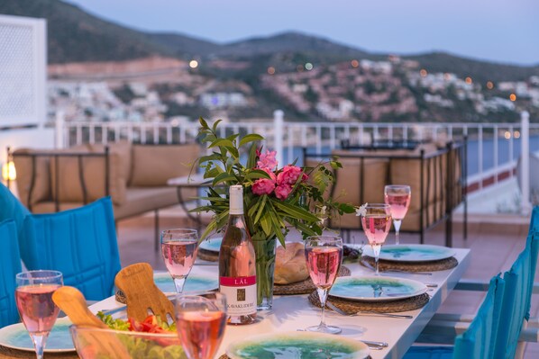 A dining table set with plates, glasses, and a bottle of wine, with a view of a coastal town in the background.