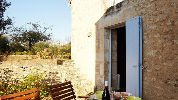 An outdoor dining area with a stone building, wooden chairs, a table set with plates, bowls, wine glasses, and a bottle.