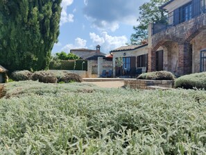 Un giardino con piante di lavanda, un vialetto in pietra e un edificio con facciata in mattoni e tetto in tegole rosse.