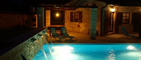 A swimming pool with lounge chairs, a stone building with a stone wall, and a night sky.