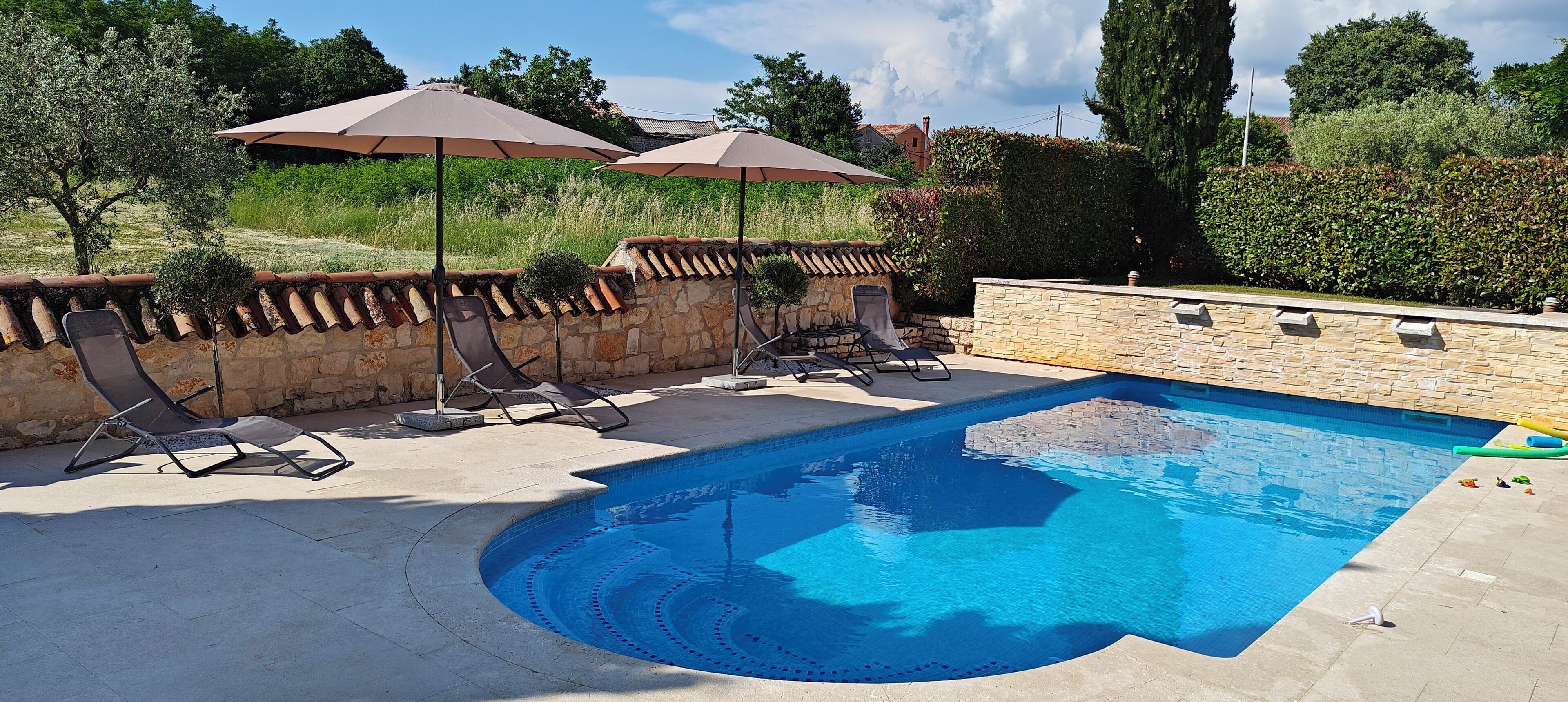 A swimming pool with lounge chairs and umbrellas, surrounded by stone walls and greenery.