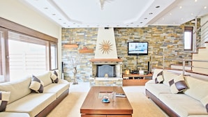 A modern living room with a stone fireplace, comfortable sofas, a coffee table, and a TV mounted on the wall. - Acharavi Beach Villa - Luxury Seaside Villa Rental in Acharavi, Island of Corfu. (Acharavi)