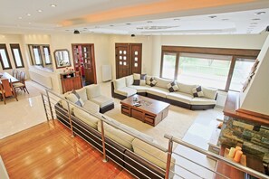 Un salon spacieux avec un grand canapé d’angle, un coin repas comprenant une longue table et des chaises, et une vue sur l’extérieur grâce à de grandes fenêtres.