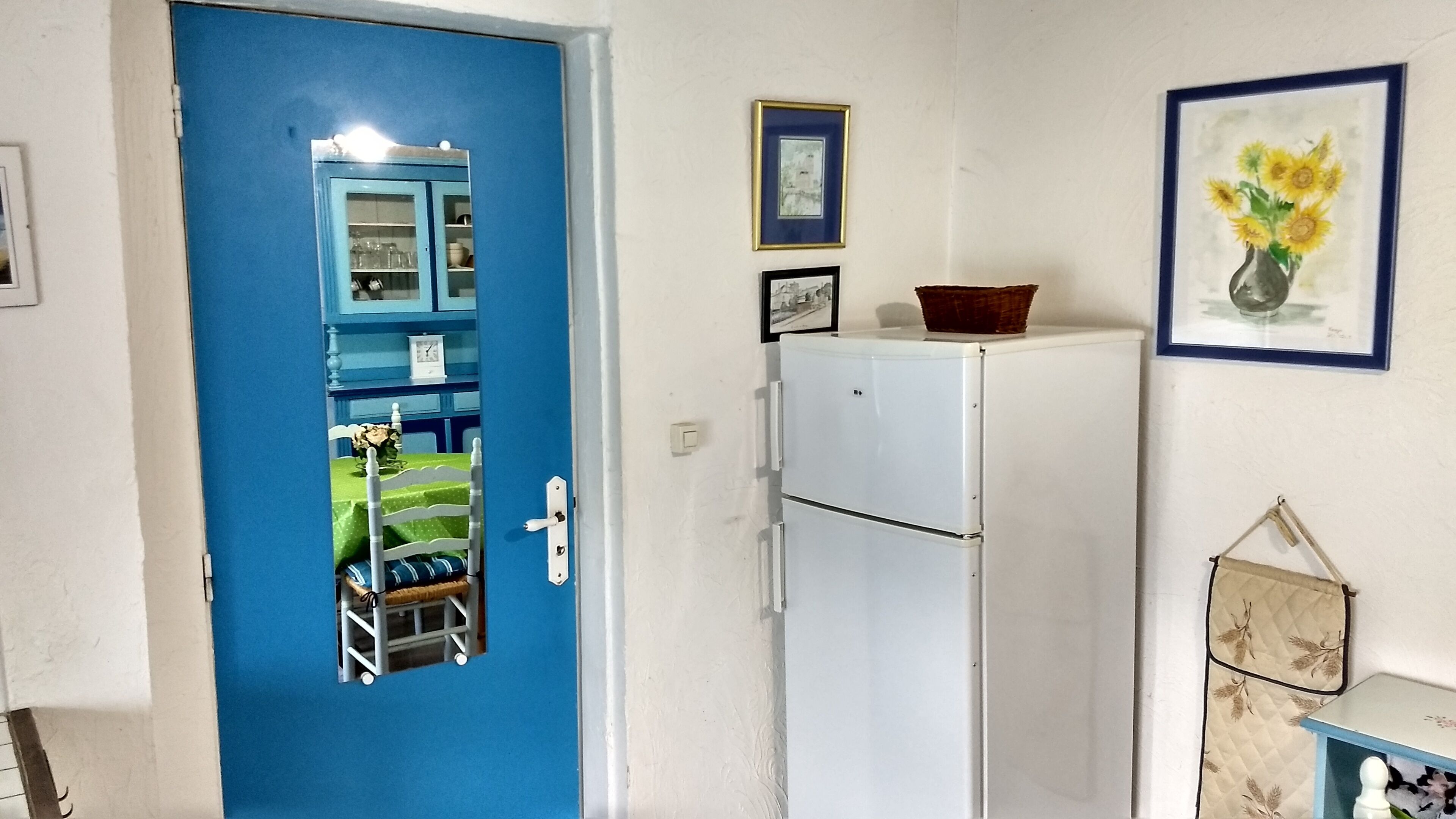 Una habitación con una puerta azul, un refrigerador blanco, una mesa pequeña con un jarrón de girasoles y un cuadro en la pared.