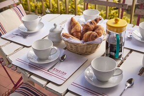 Une table dressée avec des tasses et des soucoupes blanches, un panier de croissants, un thermos et une enseigne indiquant une marque de maisons de vacances.