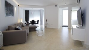 Ein modernes Wohnzimmer mit einem Ecksofa, einem Glastisch, einem Flachbildfernseher und einem Essbereich mit einer Chandelier.