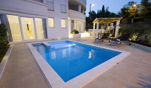 Ein rechteckiger Pool mit Fliesenrand, umgeben von einer gepflasterten Terrasse. Zwei Liegestühle mit Kissen, und ein kleiner überdachter Sitzbereich mit Tisch und Stühlen.