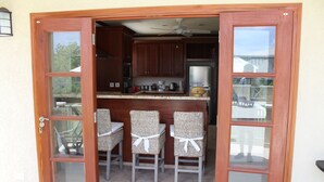 A kitchen with wooden cabinets, a countertop, and a ceiling fan. There are three wicker chairs with white ribbons tied around them. - 4 Bed Colonial Style Villa with pool in private setting, short walk to 2 beaches (Gibbes)