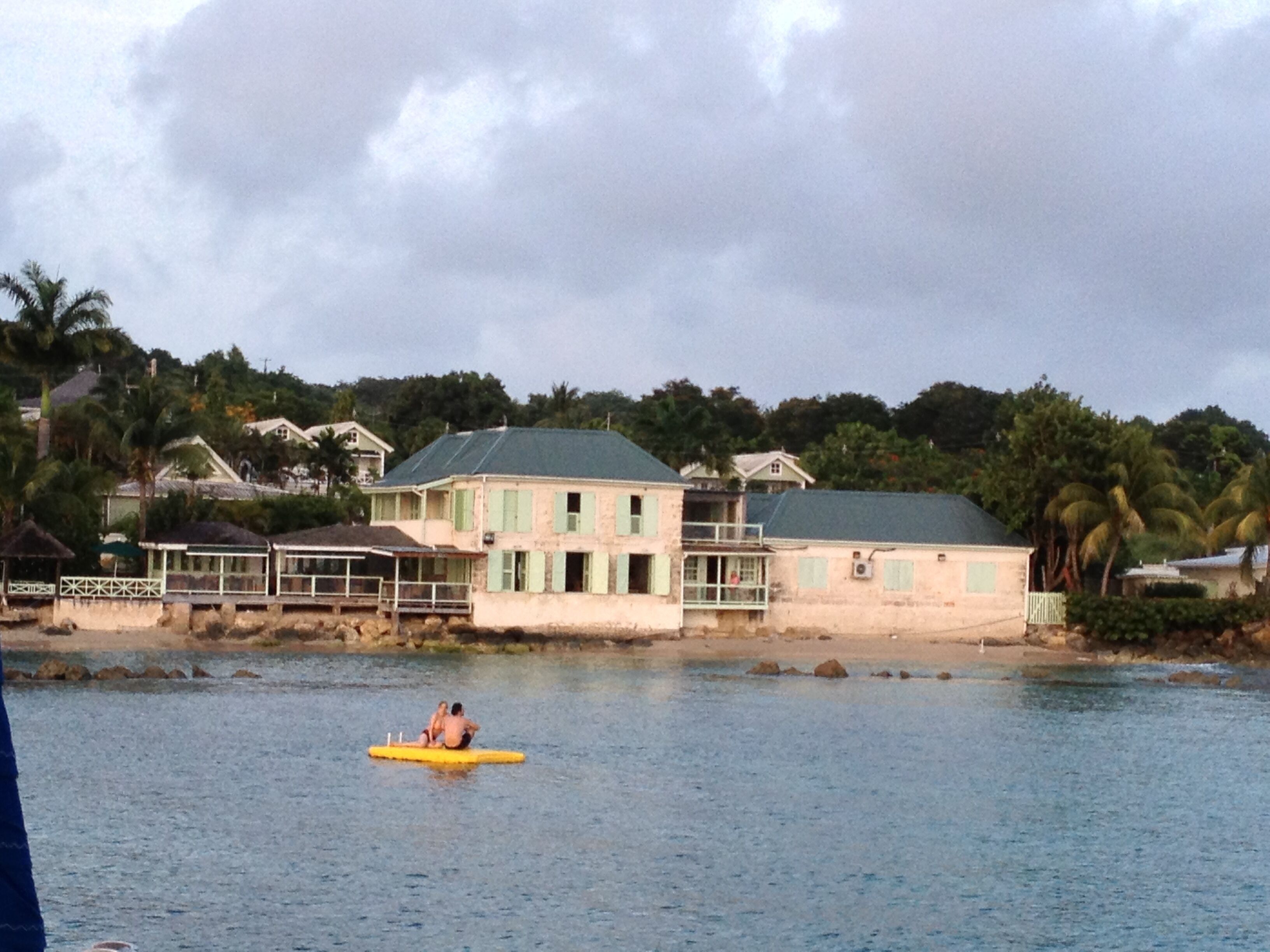 A coastal building with a balcony overlooking the sea, a person in a yellow kayak, and palm trees in the background.