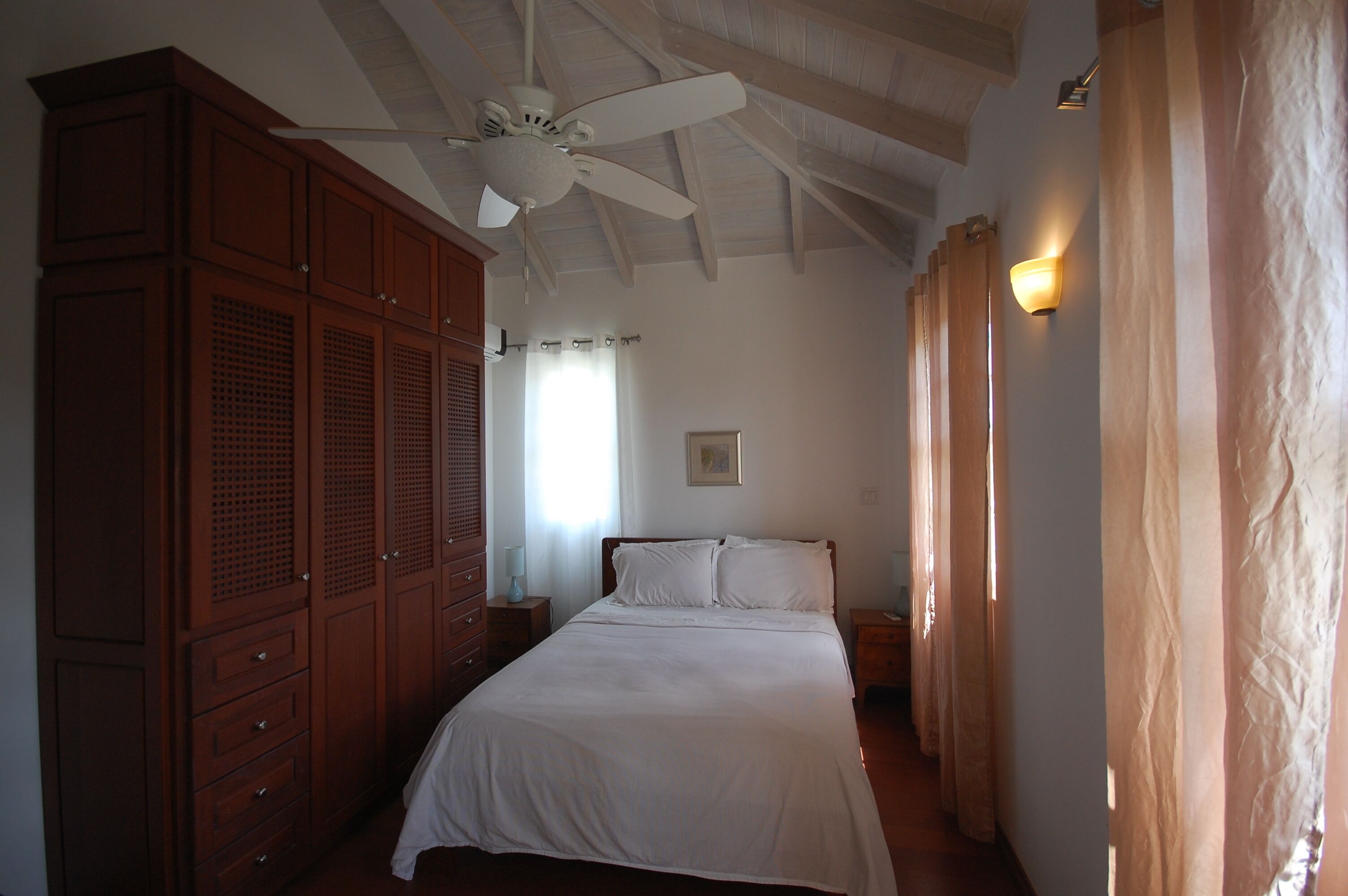 A bedroom with a bed, wooden wardrobe, ceiling fan, and a nightstand with a lamp.