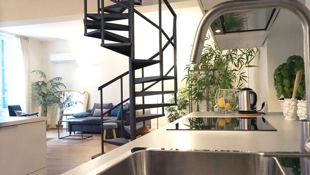 Una cocina moderna con un lavamanos de acero inoxidable, una escalera en espiral negra y una zona de comedor con sofá y plantas.