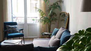 Una sala de estar con un sillón azul, un sofá con varios cojines, una planta grande en maceta y un espejo en la pared.