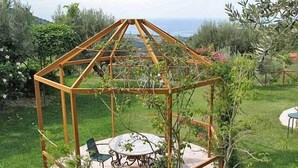 Eine hölzerne Pergola mit Tisch und Stühlen, umgeben von Grünpflanzen und Blick auf das Meer.