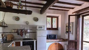 Cucina rustica con frigorifero bianco, forno a microonde e fornello. Pentole e padelle appese, e una porta in legno che dà su un giardino.