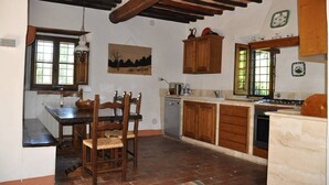 Una cuina amb armaris de fusta, una taula de dinar amb caires, una làmpada penjant i una finestra que mostra paisatge exterior.