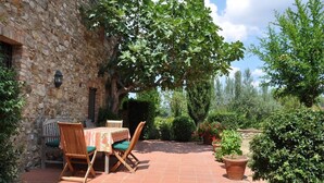 Edifici de pedra amb un pati que disposa d'una taula i cadires, envoltat de vegetació i plantes en testos.