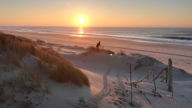 Une plage où une personne se promène le long du rivage, un coucher de soleil en arrière-plan et des dunes de sable parsemées d'empreintes.
