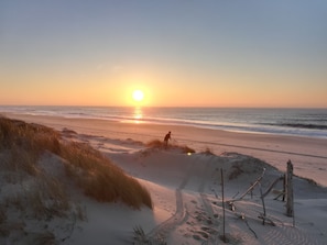 Una playa con una persona caminando por la orilla, un atardecer de fondo y dunas de arena con huellas.