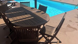 Un espace aménagé au bord de la piscine, avec une table et des chaises en bois, un coin salon abrité et une vue sur une maison moderne en arrière-plan.